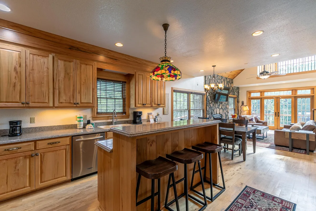 A view from the kitchen area looking into the open living area, featuring a kitchen island with a stained glass pendant light, and a glimpse of the stone fireplace and large windows beyond. The space is designed for both cooking and socializing, emphasizing the home's open-plan concept.