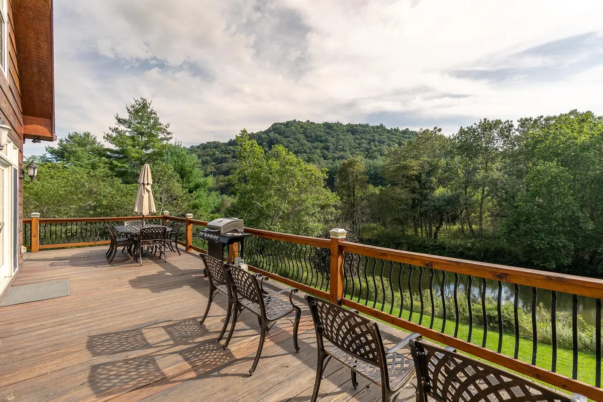 A spacious wooden deck overlooks a tranquil mountain river, with comfortable seating, a grill, and a backdrop of lush greenery. The open area offers both panoramic views of the mountains and immediate access to the outdoors, ideal for relaxation or outdoor activities.