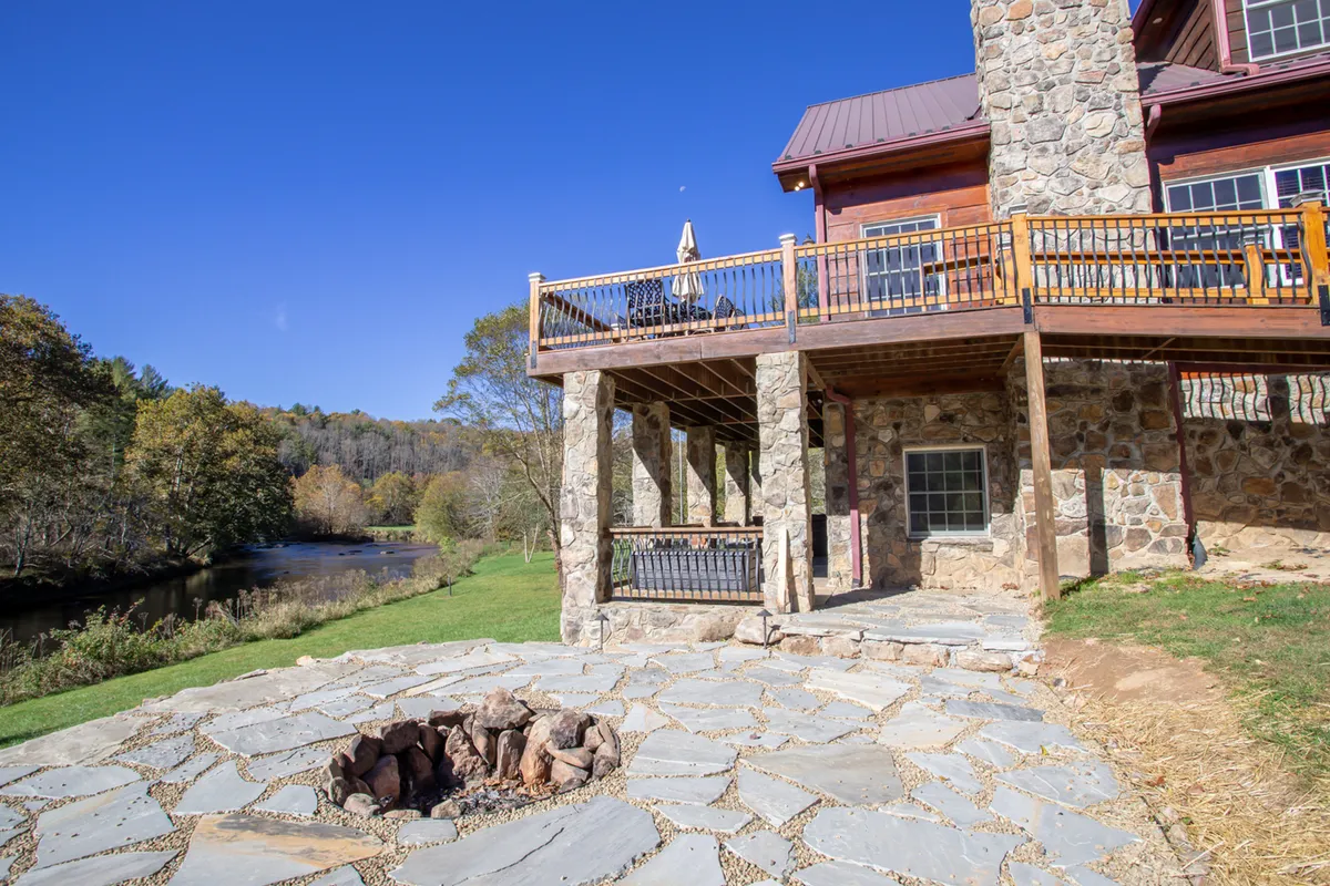 A perspective from the wooden deck shows a stone fire pit area with a direct river view, surrounded by trees. The setup demonstrates seamless flow from deck to outdoor gathering spaces and natural elements, perfectly capturing the resort-like playground vibe.