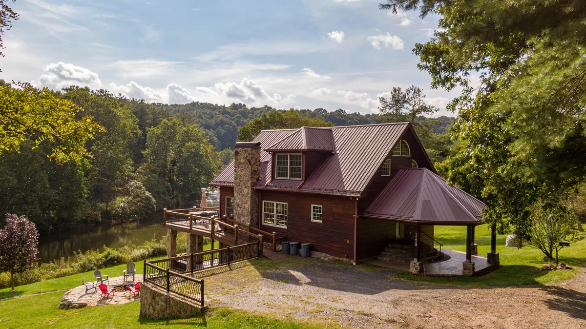 A picturesque wooden cabin with a stone chimney and a deck overlooking the water. The setting is lush, with greenery surrounding the cabin, and the deck offers an inviting space for relaxation or social gatherings by the river.