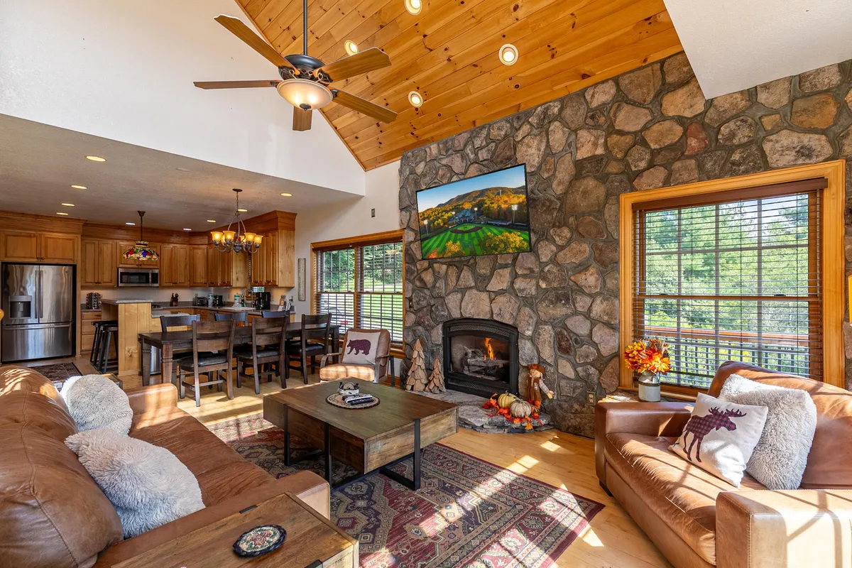 A wide, open-plan living and dining area with a soaring wooden ceiling and a striking stone fireplace. Comfortable seating is arranged around the fireplace, and large windows flood the space with natural light while framing views of the river and mountains. The dining area sits adjacent, creating a seamless flow for gatherings and meals.