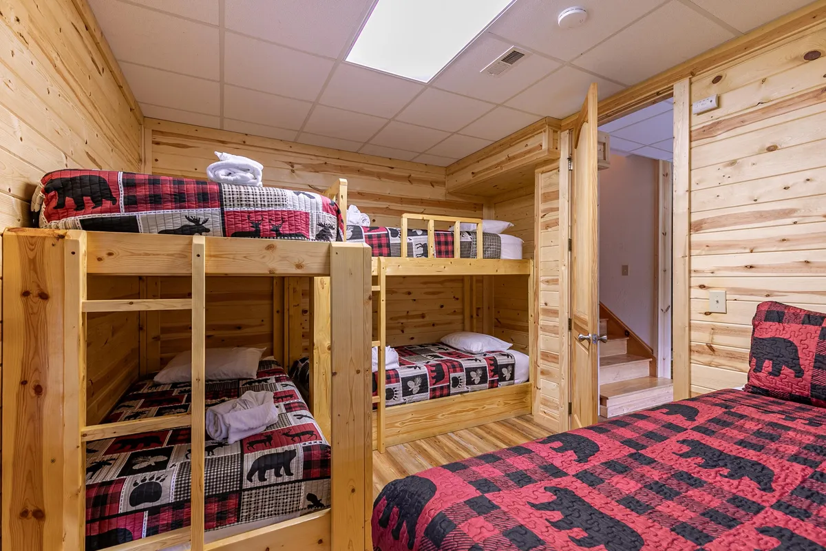 A cheerful bunk room styled with wood paneling and bear-patterned bedding, featuring multiple bunk beds and a single bed. The inviting mood is perfect for kids, cousins, or friends looking for a playful sleepover experience.