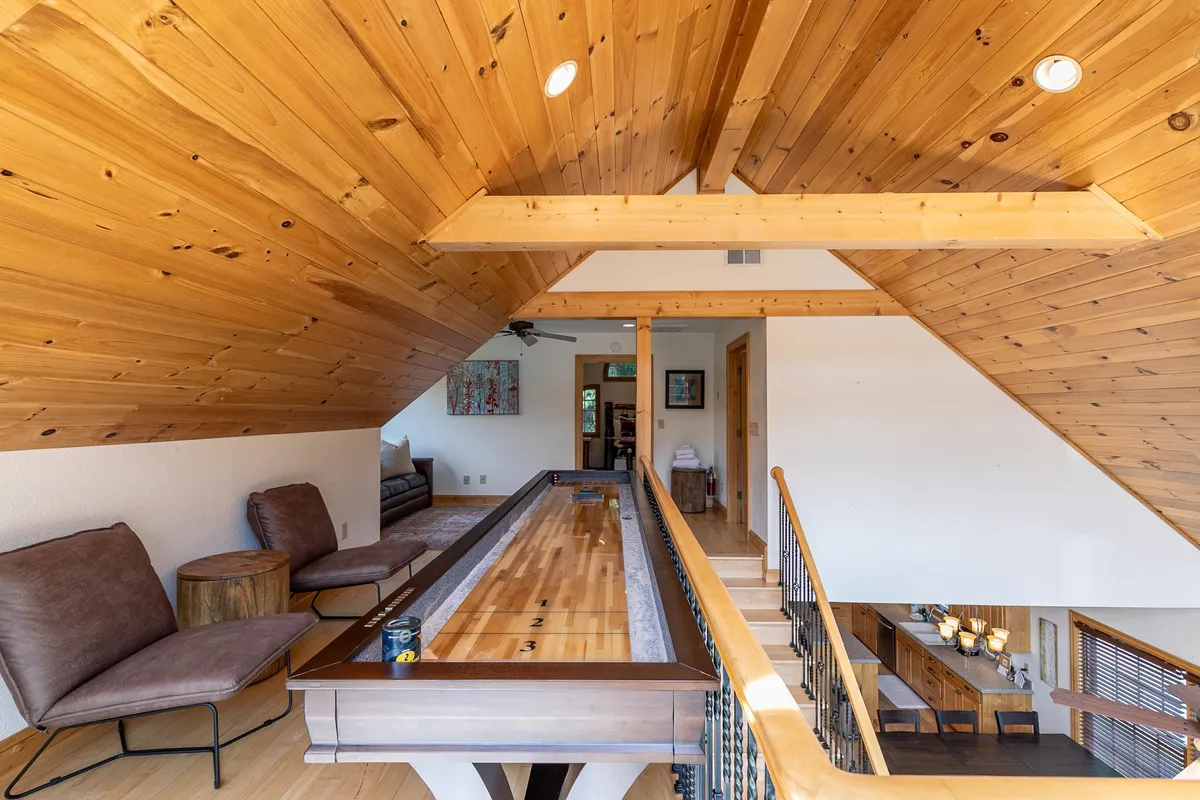 A cozy loft area featuring a shuffleboard table, inviting seating beneath a wooden ceiling, and soft lighting that encourages playful competition and laid-back relaxation. The scene is intimate and playful, ideal for casual gatherings or family game nights.
