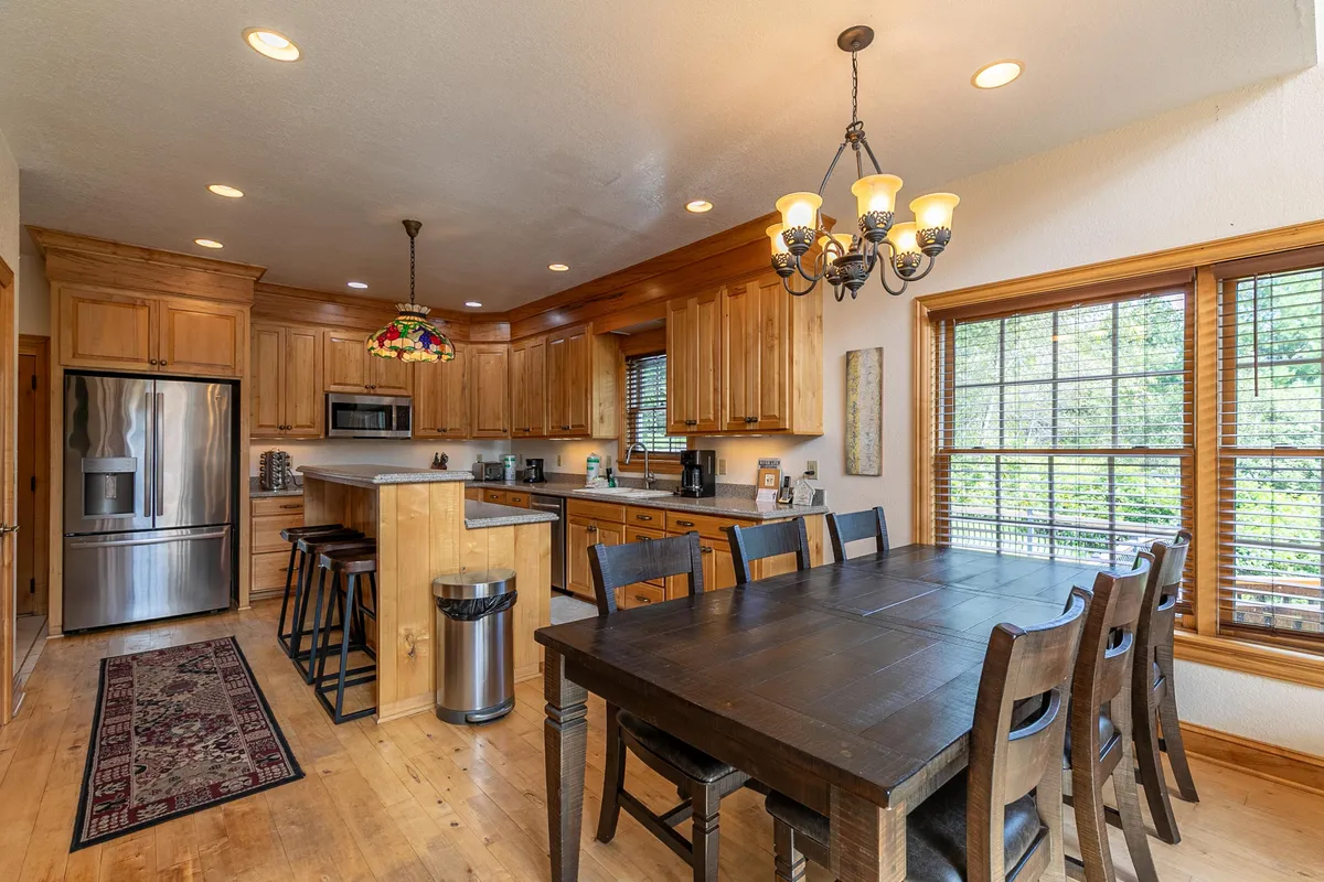 A spacious dining area featuring a large wooden table, stainless steel appliances, and a colorful chandelier. The setting is bright, inviting, and ready for family meals or entertaining, with clear views into the kitchen and hints of natural light from nearby windows.