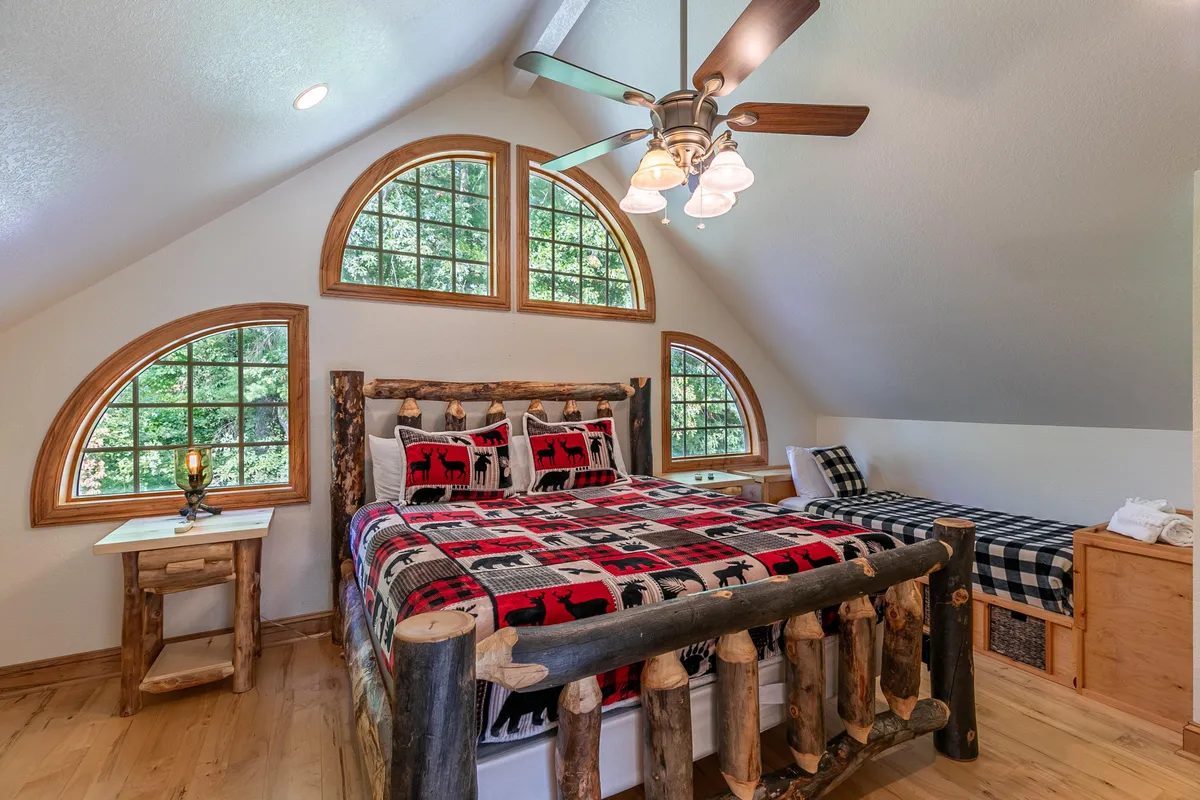 A warm, rustic bedroom with a log bed featuring patterned bedding, arched windows overlooking the outdoors, a wooden bedside table, and an additional bed with a cozy checkered blanket. The space feels inviting and ideal for family stays.