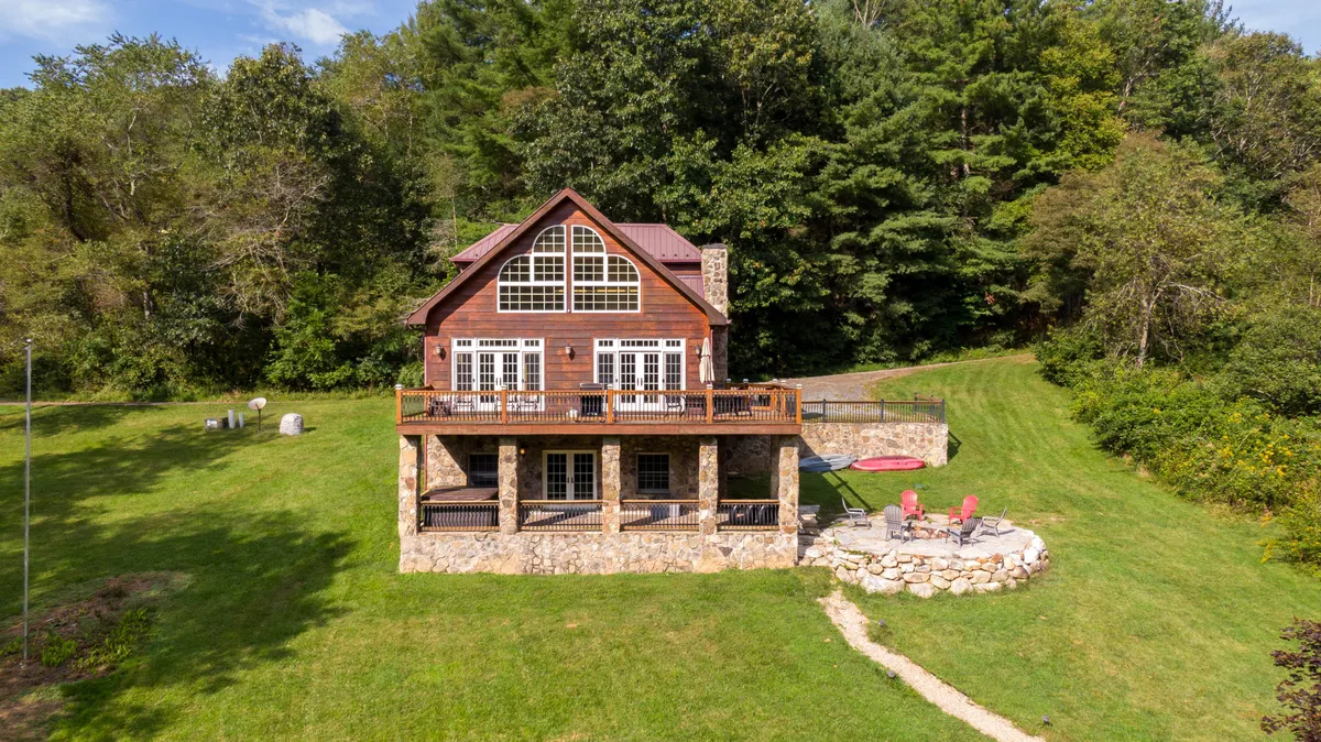 A house with large windows sits beside a green lawn surrounded by trees. The stone patio provides a transition from indoor to outdoor living, and the spacious lawn is ideal for playful activities, making it perfect for pets to roam and families to gather.