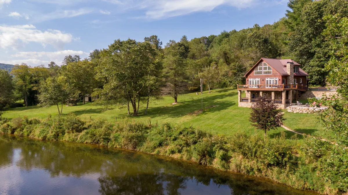 A wide-angle exterior view of the house set directly on the riverbank, with the New River flowing gently beside the home. Large trees provide shade, and the lush landscape creates an inviting path to the water's edge, illustrating direct river access right from the property.