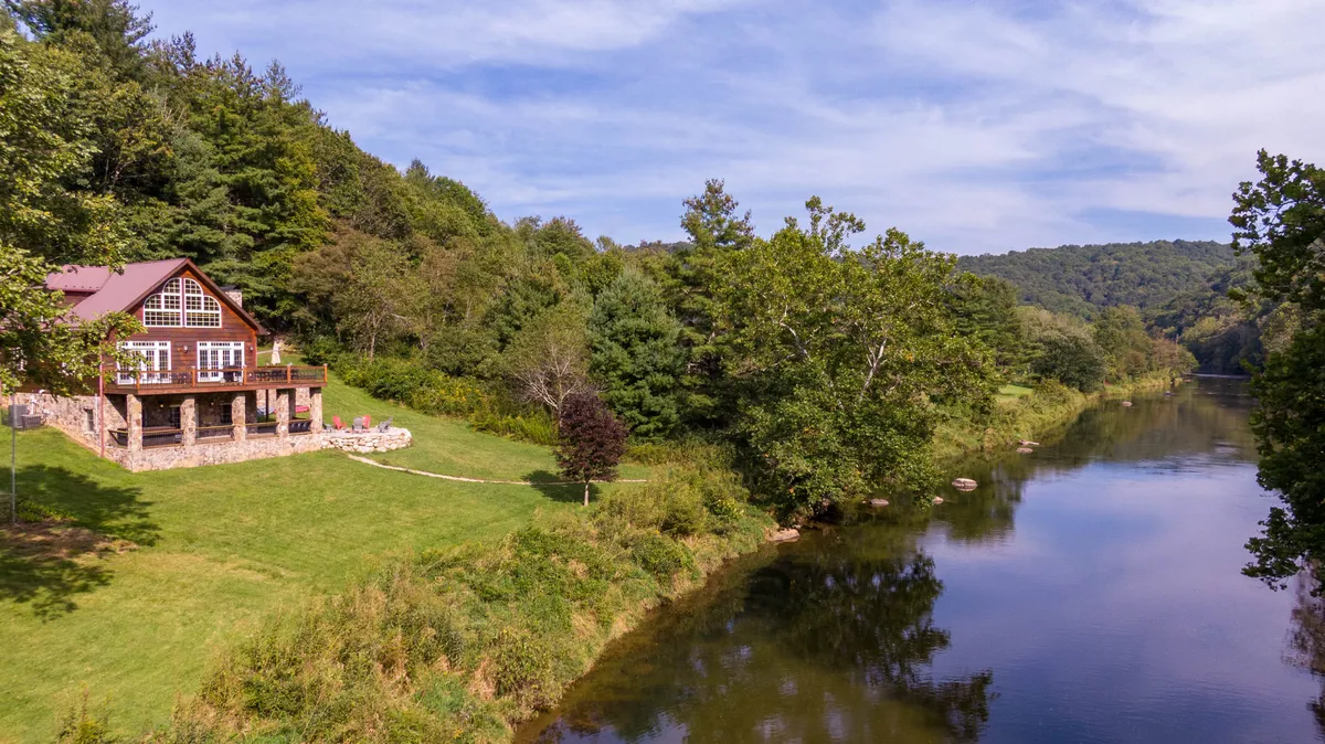 A wide view reveals the lush, expansive lawn stretching down to the river’s edge, with the house, trees, and flowing water all visible. The space is inviting for activities like frisbee, fetch, picnics, and riverside lounging, as described in the section.