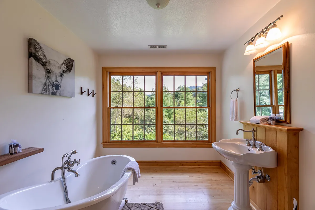 A luxurious bathroom with a freestanding bathtub positioned by large windows offering a scenic view, double sinks, wooden accents, and deer artwork. The tranquil setting invites relaxation and effortless routines.
