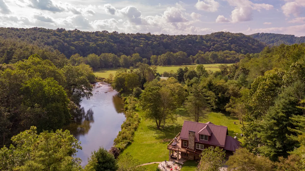 A peaceful riverfront mountain house nestled among tall trees, with the serene New River flowing beside a spacious grassy yard. The sunset casts a golden glow over the river, inviting outdoor activities like fishing, picnicking, or simply relaxing by the water. The home's inviting exterior promises comfort and scenic views.