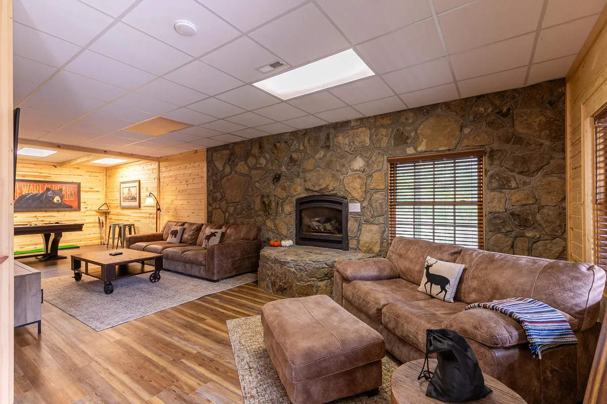A spacious, inviting lower-level den with a large sectional sofa gathered around a stone fireplace and a pool table. The setting is designed for relaxation and group activities, with warm lighting and rustic touches creating the perfect ambiance for tournament nights or movie marathons.