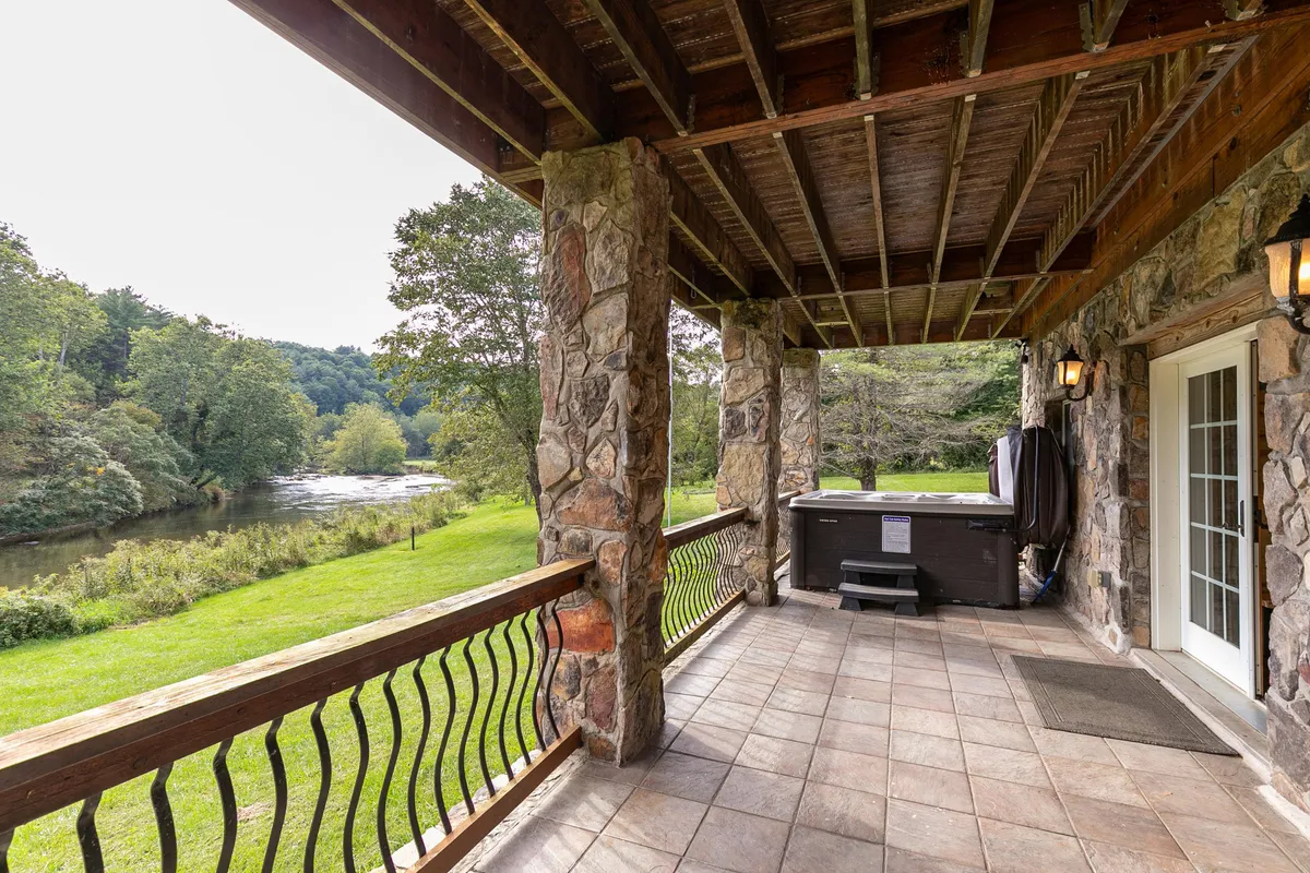 This image captures an inviting exterior space featuring a stone patio with a hot tub overlooking a scenic river. There are stone pillars framing the scene, with lush greenery and the river in clear view, conveying a serene, relaxing atmosphere for outdoor enjoyment.