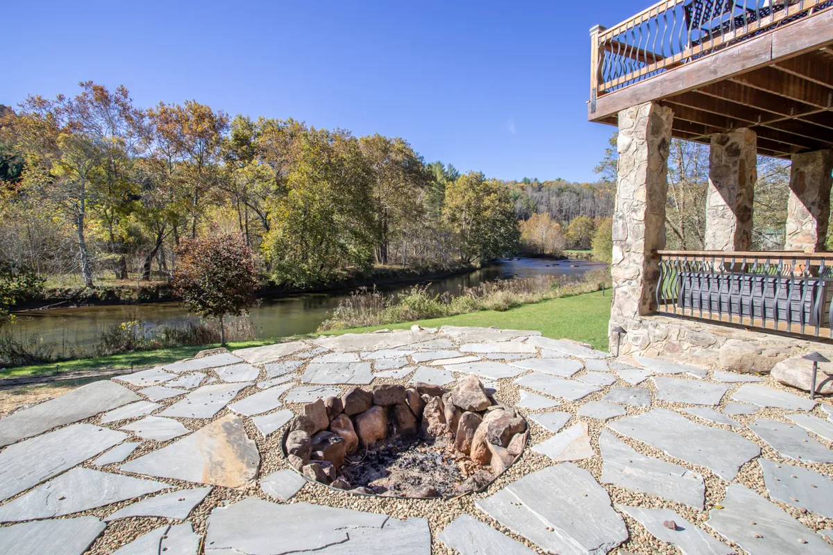 An exterior scene featuring a cozy firepit area near the river, with mature trees and a balcony visible. The river glistens in the background, emphasizing the tranquil setting and the inviting atmosphere for riverside relaxation and group activities after a day of adventure.
