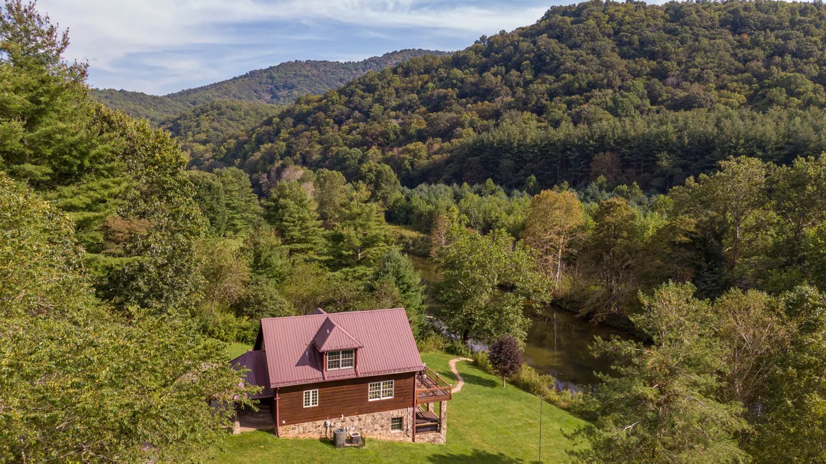 A wide shot of the cabin nestled among mature trees, with the river winding nearby and mountains in the background. The setting conveys peaceful seclusion and direct connection to nature, with the cabin serving as a cozy refuge for all seasons.