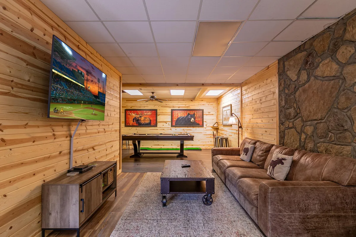 A fun, casual interior game room with a pool table, large screen TV, comfortable seating, and stone wall accents. The cozy setup suggests a lively, social space for family and friends to play games or unwind indoors after a day outside.